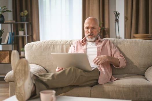 Senior man with distinguished silver beard depicted enjoying time home. He seated comfortably on sofa, engrossed in watching internet on his laptop. High quality photo