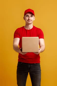 Delivery man employee in red cap, red t-shirt holding cardboard box isolated on yellow background