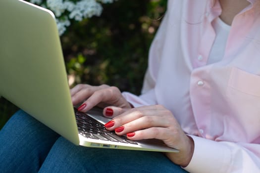 A girl in a pink shirt sits in the garden on the grass with a laptop, hands with a red manicure