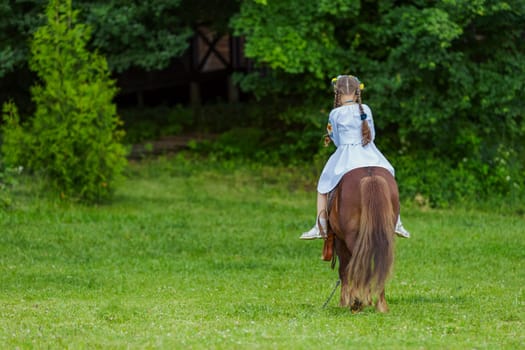 A little girl in the Ukrainian national costume rides a pony on the lawn