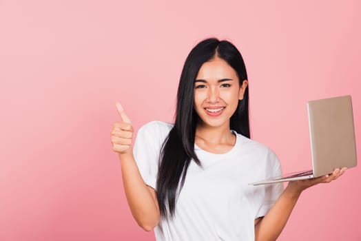 Portrait of happy Asian beautiful young woman confident smiling holding using laptop computer and showing thumb up for like gesture, studio shot isolated on pink background, with copy space