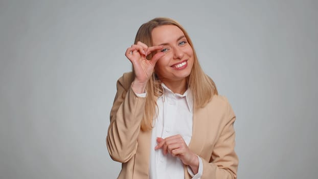 Need some more, please give me. Woman in formal suit showing a little bit gesture with sceptic smile, showing minimum sign, measuring small size. Business girl isolated alone on gray studio background