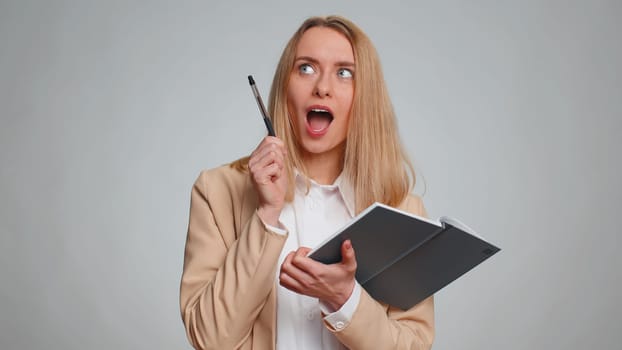 Thoughtful journalist young businesswoman making notes, writing down thoughts with pen into notepad notebook diary, to do list, good idea. Female girl in formal suit. Woman isolated on gray background