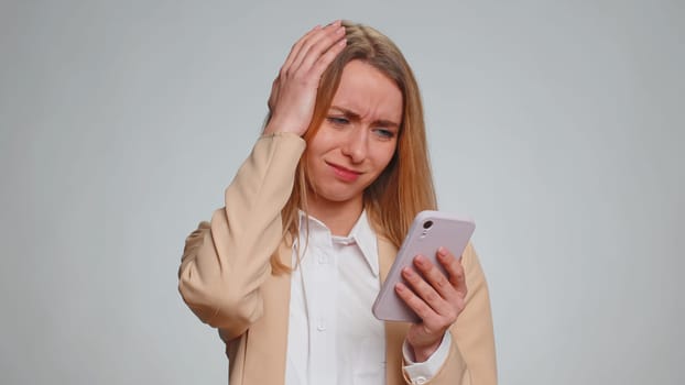 Sad business woman use mobile cell phone typing browsing, loses becoming surprised sudden lottery results, bad fortune, loss, fail. Unhappy girl isolated alone on gray studio wall background, indoors