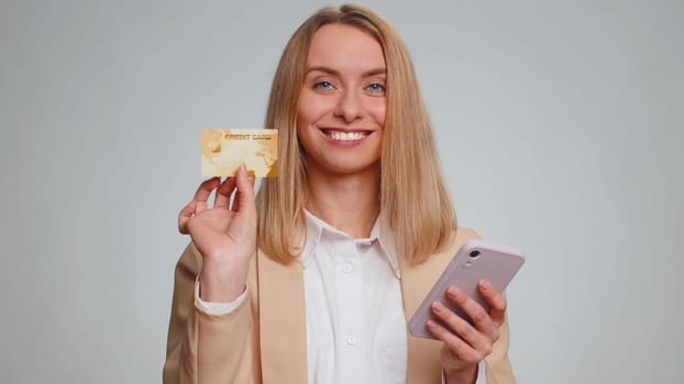 Sincere business woman customer using credit bank card and smartphone while transferring money, purchases online shopping, payment. Finance and internet. Young girl isolated alone on gray background