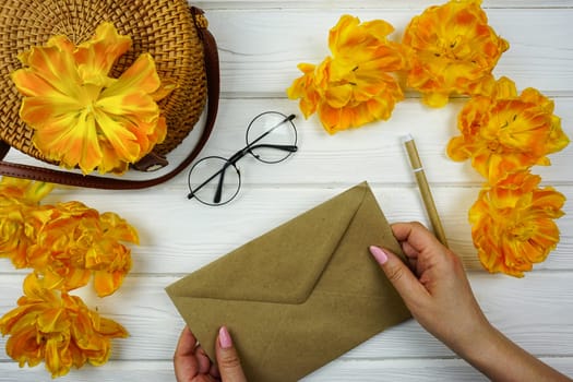 Women's hands are holding a craft envelope. On a white wooden table there is a wicker bag and yellow terry tulips.