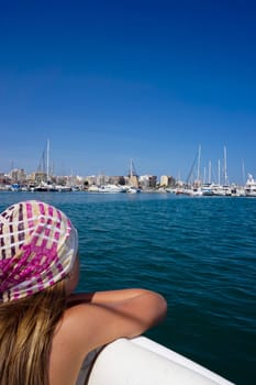 A tanned girl looks from a boat at the blue sea. City buildings can be seen in the distance.