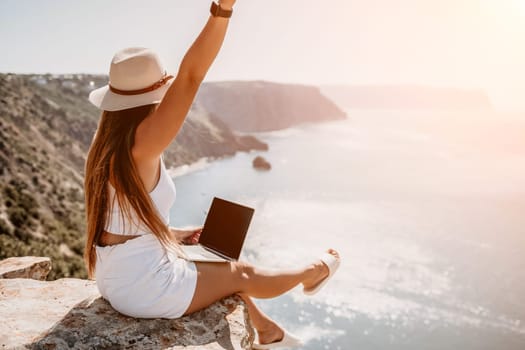 Successful business woman in yellow hat working on laptop by the sea. Pretty lady typing on computer at summer day outdoors. Freelance, travel and holidays concept.