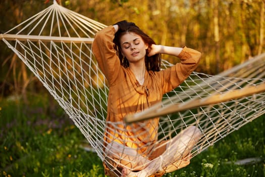 a joyful woman is sitting in a mesh hammock in nature, relaxing and enjoying the rays of the setting sun, straightening her hair with her hands, closing her eyes on a warm summer day. Horizontal photo on the theme of outdoor recreation. High quality photo