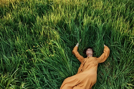 a sweet, calm woman in an orange dress lies in a green field with her arms outstretched, enjoying the silence and peace. Horizontal photo taken from above. High quality photo