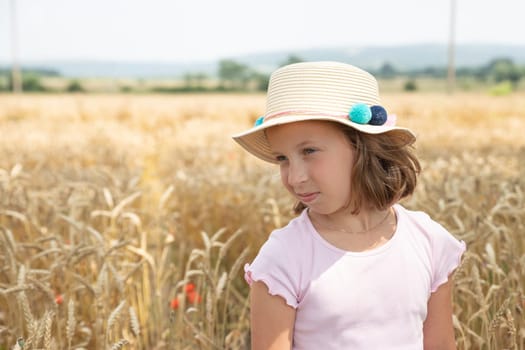 Adorable little blonde girl in a hat and a white linen dress, standing on a wheat field and touching the ears of wheat with her hands.