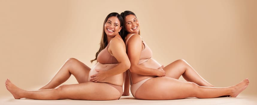 Pregnant woman friends, studio and underwear with smile, sitting and together for support by backdrop. Happy women, pregnancy and solidarity for wellness, care and holding healthy stomach by backdrop.