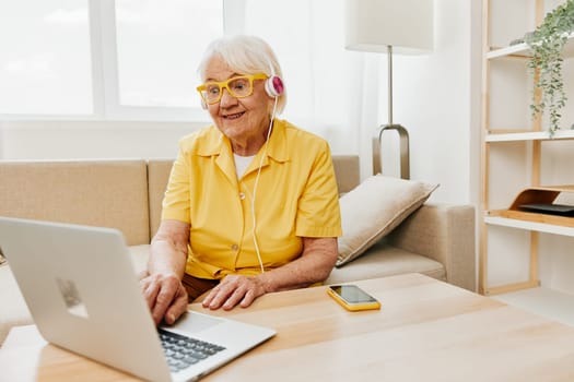 Happy elderly woman with a laptop typing in headphones smile sitting at home on the couch in a yellow shirt, bright modern interior, lifestyle online communication. High quality photo