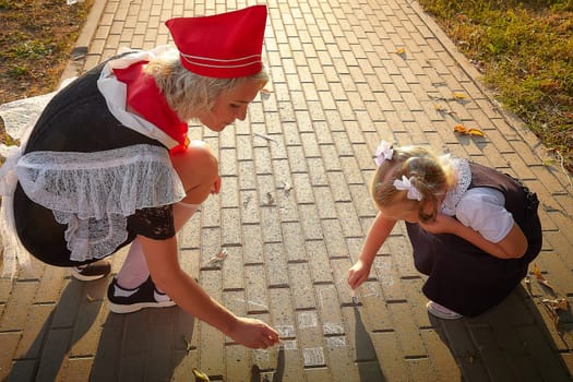 Young and adult schoolgirl on September 1 drawing by chalk on asphalt. Generation of schoolchildren of USSR and Russia. Female pioneer in red tie and October girl in modern uniform. Daughter and mom