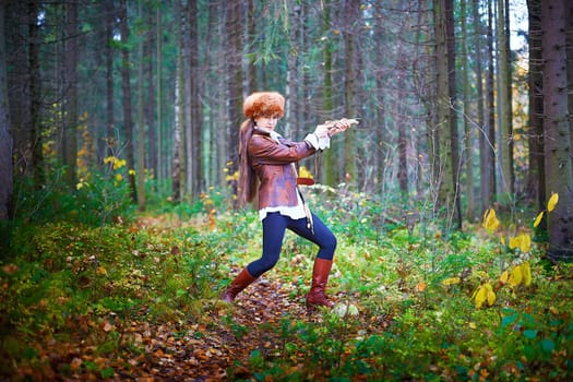 Girl in a leather jacket, a big red fox fur hat and with a crossbow in the forest in autumn. A female model poses as a fabulous royal huntress on nature hunt at a photo shoot