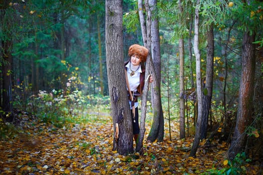 Girl in a leather jacket, a big red fox fur hat and with a crossbow in the forest in autumn. A female model poses as a fabulous royal huntress on nature hunt at a photo shoot