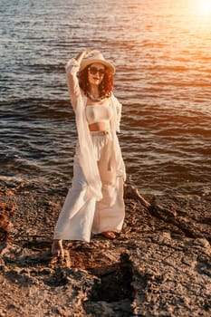 woman sea sunset. woman in a white pantsuit and hat is standing on the beach enjoying the sea. Happy summer holidays.