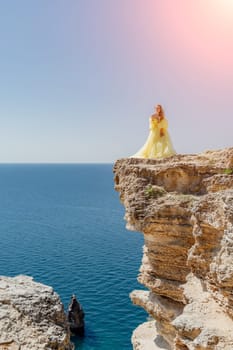 Woman yellow dress sea. Side view Young beautiful sensual woman in yellow long dress posing on a rock high above the sea at sunset. Girl in nature against the blue sky.