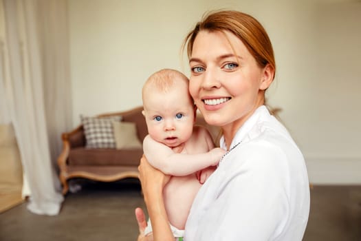 Cheerful mother in white robe smiling and looking at camera while embracing adorable baby at home against blurred background