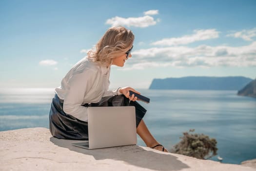 Freelance women sea working on a computer. Pretty middle aged woman with computer and phone outdoors with beautiful sea view. The concept of remote work