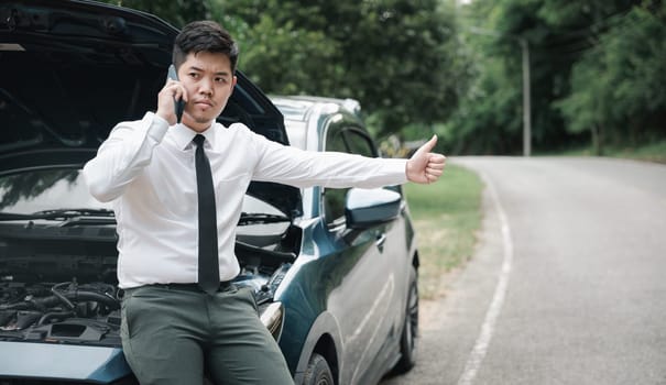 Asian businessman stranded on roadside after car breakdown, using phone to call for help. Candid shot of man waiting for assistance and thumbing up. Transportation and technology concept.