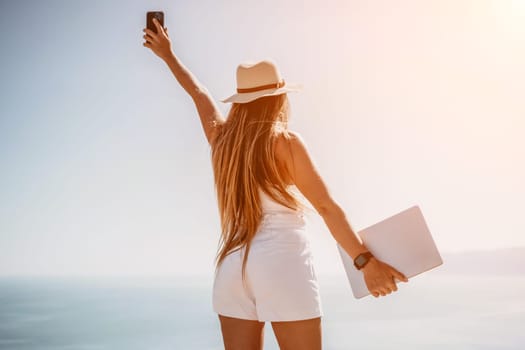 Successful business woman in yellow hat working on laptop by the sea. Pretty lady typing on computer at summer day outdoors. Freelance, travel and holidays concept.