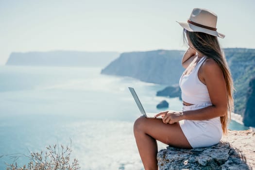 Successful business woman in yellow hat working on laptop by the sea. Pretty lady typing on computer at summer day outdoors. Freelance, travel and holidays concept.