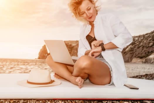 Successful business woman in yellow hat working on laptop by the sea. Pretty lady typing on computer at summer day outdoors. Freelance, travel and holidays concept.