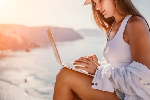 Digital nomad, Business woman working on laptop by the sea. Pretty lady typing on computer by the sea at sunset, makes a business transaction online from a distance. Freelance remote work on vacation