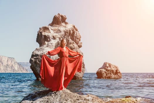 Woman travel sea. Young Happy woman in a long red dress posing on a beach near the sea on background of volcanic rocks, like in Iceland, sharing travel adventure journey