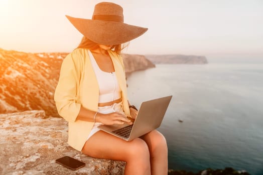 Successful business woman in yellow hat working on laptop by the sea. Pretty lady typing on computer at summer day outdoors. Freelance, travel and holidays concept.