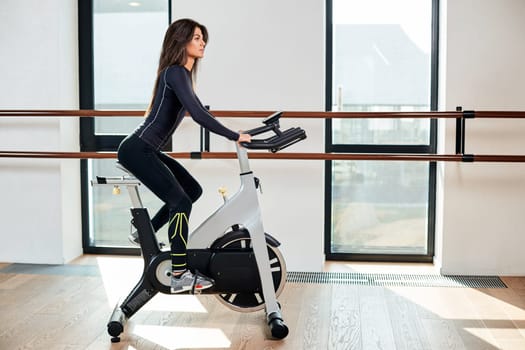 Muscular young woman working out on the exercise bike at the gym, intense cardio workout. She is smiling and looking ahead
