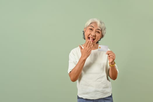 Excited senior woman with credit card isolated on green pastel background. Shopping and payment concept.