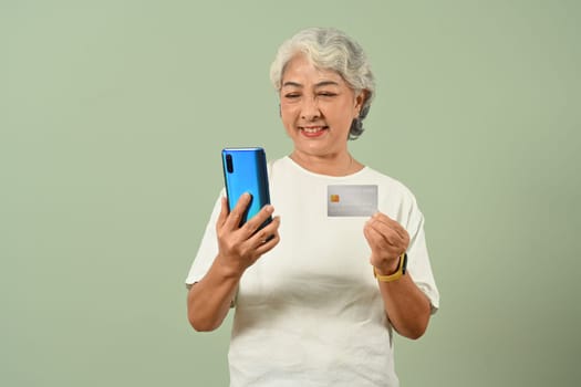 Satisfied mature woman 50s years with credit card using mobile phone to ordering in internet, standing over green pastel background.