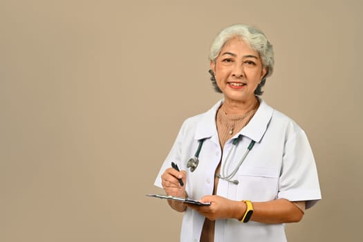 Confident mature doctor wearing white medical overall and stethoscope standing on beige background. Medical, healthcare concept.