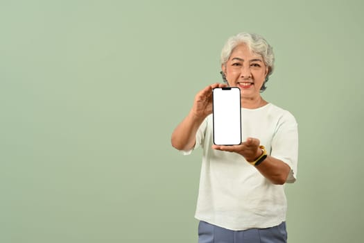 Positive senior gray haired woman showing smartphone with blank screen isolated on green background with space for text.