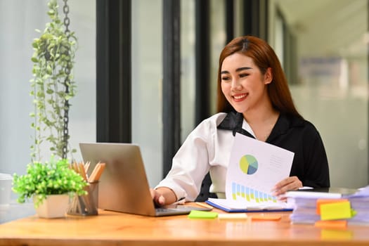 Image of cheerful asian female office worker reading email, using laptop on wooden desk. Business and technology concept.