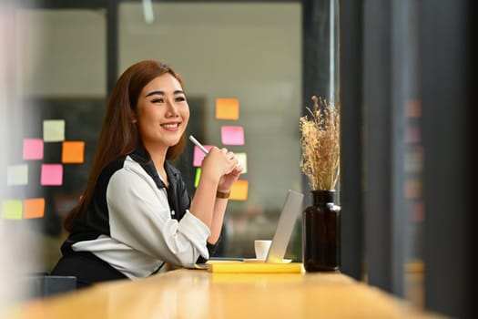 Smiling millennial woman entrepreneur sitting at wooden counter in office and looking through window.