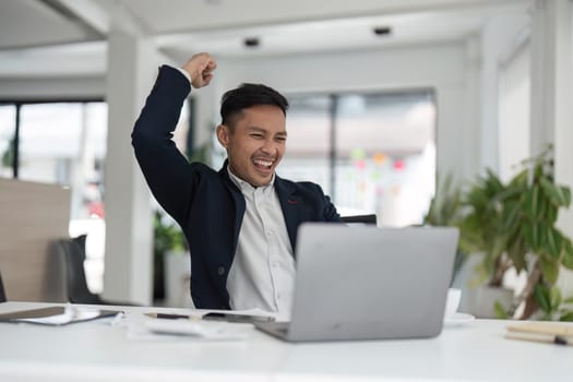 Happy young businessman looking at laptop excited by good news online, lucky successful winner man sitting at office desk raising hand in yes gesture celebrating business success win result.