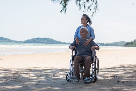 Happy Asian senior man on wheelchair and elderly woman couple smiling on sand of the beach for taking the patient travel together