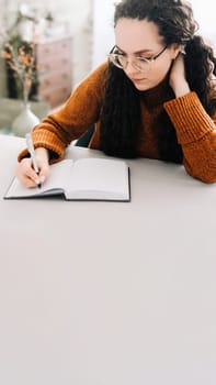 Young woman multitasking: writing in journal and studying for success. Close-up view of woman writing in copybook for studying or making checklist. Focused young woman taking notes in paper notebook.