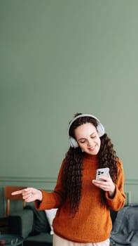 Happy pretty young woman wearing headphones using a smartphone dancing at home. Cheerful girl listening music on mobile phone, singing song feeling relaxed standing in modern living room interior