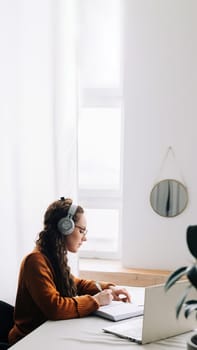 Young student attending virtual seminar while studying at home. Woman wearing headphones and typing on laptop, taking notes during e-learning session or online class