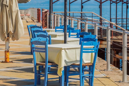 Empty tavern restaurant by the seafront with wooden chairs and tables under a bright sun in summer.