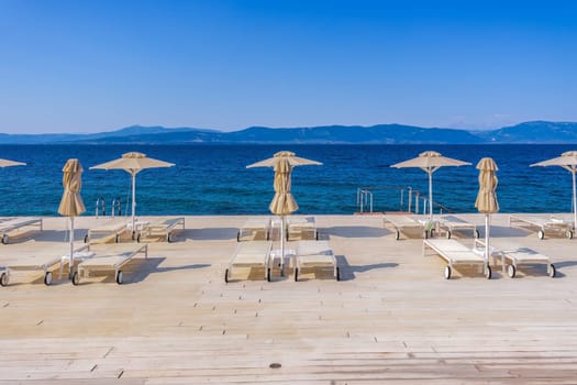 Empty wooden sunbathing deck with chairs and umbrellas by a calm blue sea under the bright sun in summer.