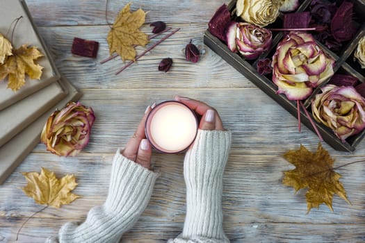 Women's hands hold a burning candle. Books in craft covers are laid out on the table, there is a wooden box with dried rose flowers and yellow autumn leaves.