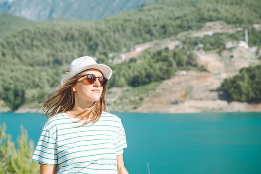 Smiling woman in hat and sunglasses with wild hair standing near mountains lake on background. Positive young woman traveling on blue lake outdoors travel adventure vacation.
