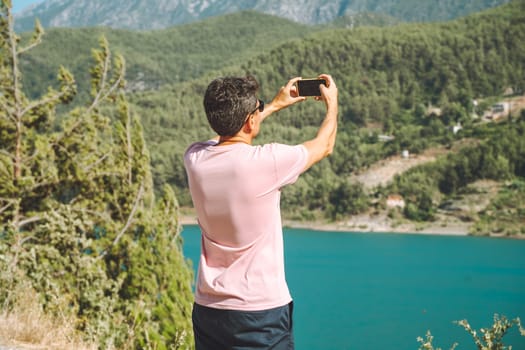 Man shooting a video on mobile phone of mountains lake background. Traveler male taking photo on cellphone on the blue lake outdoors travel adventure vacation.