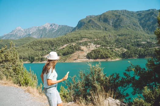 Smiling woman in hat and sunglasses with wild hair standing near mountains lake on background. Positive young woman traveling on blue lake outdoors travel adventure vacation.