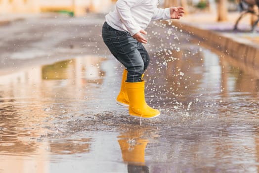 Child's feet in yellow rubber boots jumping over a puddle in the rain.
